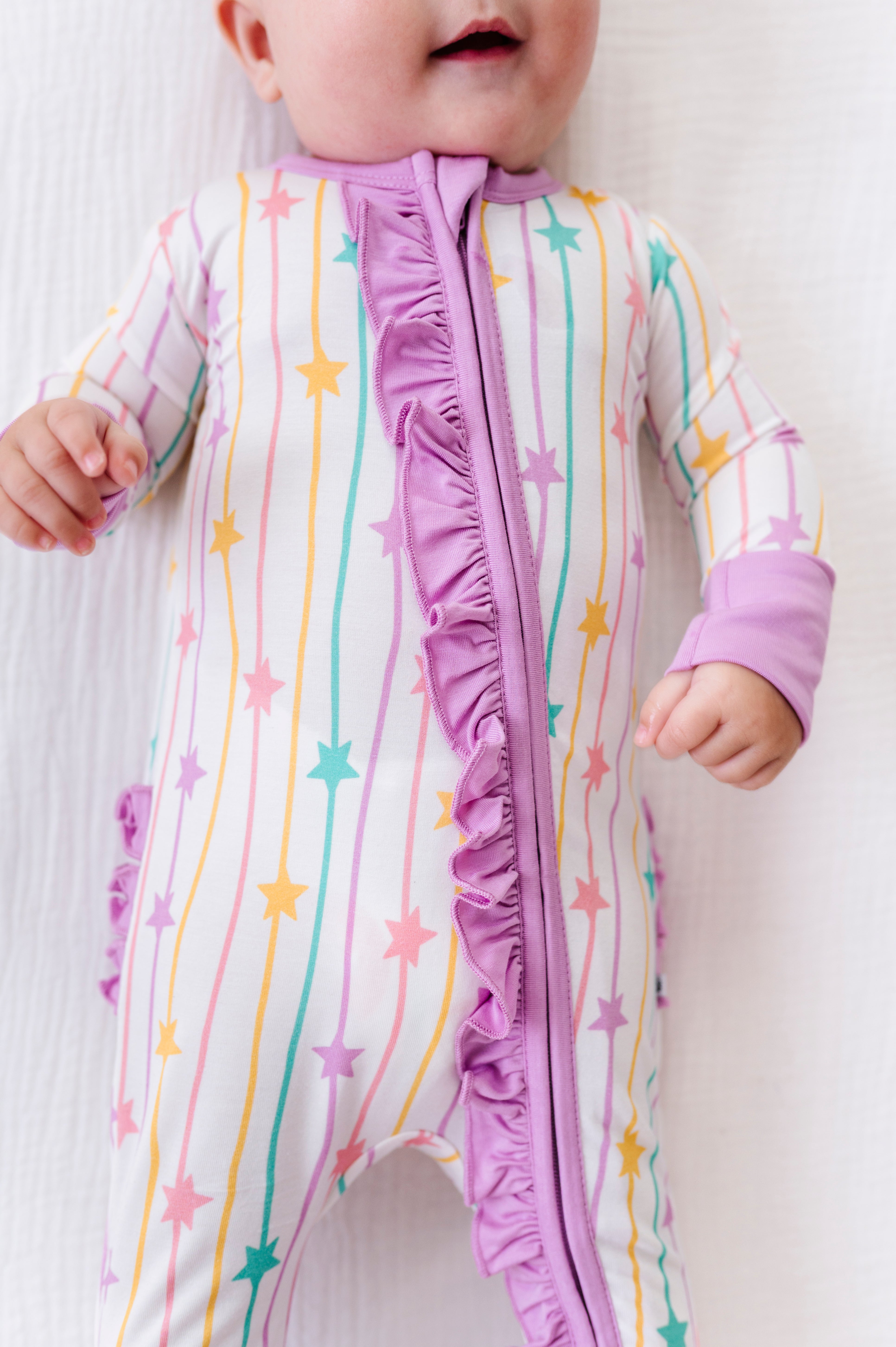A baby wearing a long-sleeve footless sleep sack with a vertical ruffle zipper. The sleep sack has a white base with vertical pastel stripes in pink, yellow, and teal, accented with pastel pink and teal stars.